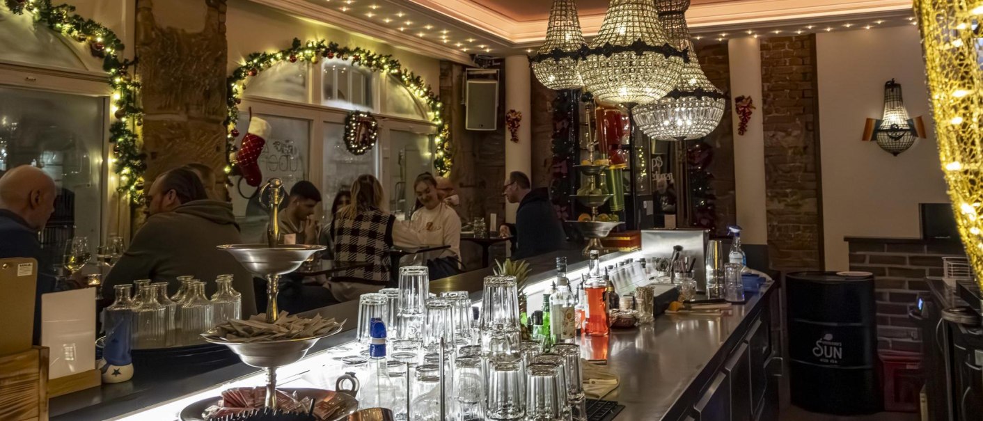 Festlich dekorierte Bar mit Kronleuchtern, Gläsern und Getränken auf der Theke. Gäste sitzen und unterhalten sich in gemütlicher Atmosphäre., © Stuttgart Marketing GmbH, Sarah Schmid