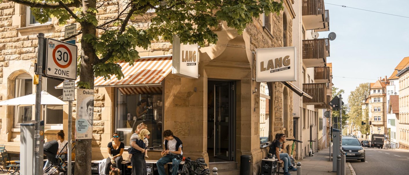 Café Lang an einer Straßenecke mit Menschen, die draußen sitzen. Ein 30er-Zone-Schild und Bäume sind ebenfalls zu sehen., © SMG, Sarah Schmid