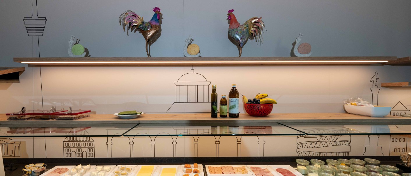 Ein reichhaltiges Frühstücksbuffet mit Käse, Aufschnitt, Gemüse und Joghurt vor einer Wand mit Stuttgart-Motiven und dekorativen Hähnen., © TOMAS Ein reichhaltiges Frühstücksbuffet mit Käse, Aufschnitt, Gemüse und Joghurt vor einer Wand mit Stuttgart-Motiven und dekorativen Hähnen., © TOMAS