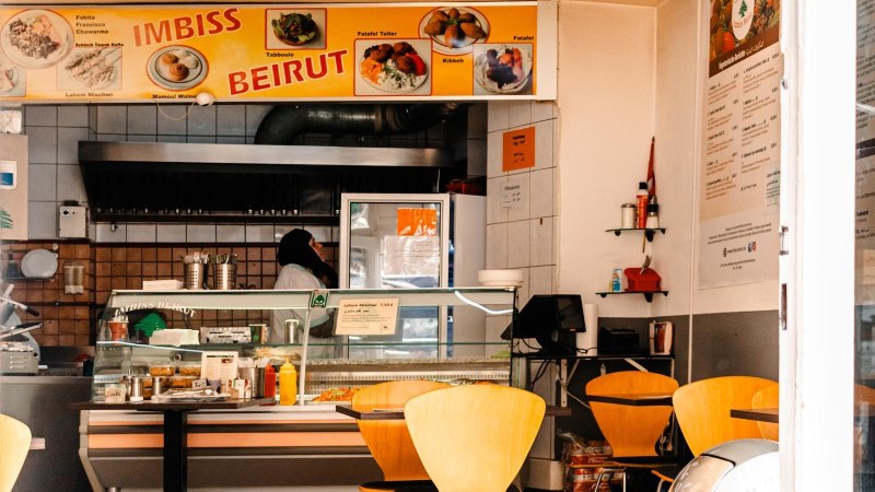 Ein Imbiss mit gelbem Schild 'Imbiss Beirut', leeren Tischen und einer Person hinter der Theke., © SMG, Sarah Schmid Ein Imbiss mit gelbem Schild 'Imbiss Beirut', leeren Tischen und einer Person hinter der Theke., © SMG, Sarah Schmid
