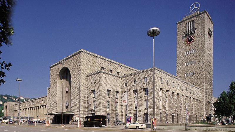 Der Hauptbahnhof Stuttgart mit seinem markanten Uhrturm und Mercedes-Stern, umgeben von Autos und Bäumen, unter einem klaren blauen Himmel., © Stuttgart-Marketing GmbH Der Hauptbahnhof Stuttgart mit seinem markanten Uhrturm und Mercedes-Stern, umgeben von Autos und Bäumen, unter einem klaren blauen Himmel., © Stuttgart-Marketing GmbH