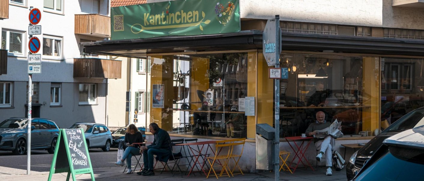 Ein Café namens Kantinchen mit gelbem und grünem Schild. Draußen sitzen Menschen an bunten Tischen. Verkehrsschilder und Autos sind zu sehen., © SMG, Sarah Schmid