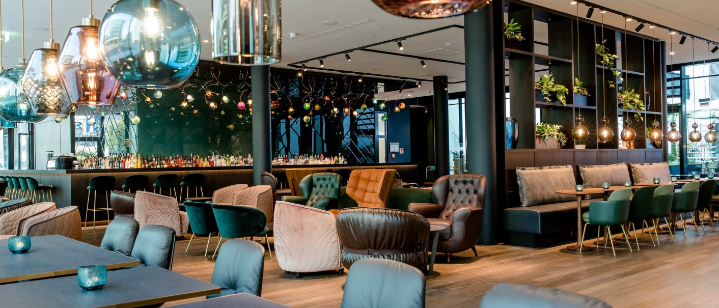 A modern restaurant with elegant hanging lamps, cozy armchairs and a bar in the background. Plants decorate the shelves., &copy; Motel One - Stuttgart Hauptbahnhof