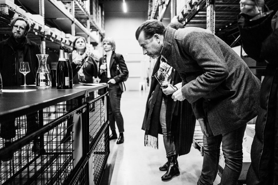 Mehrere Personen stehen in einem Lagerraum und probieren Champagner. Eine Person beugt sich vor, um ein Glas zu riechen. Schwarz-Weiß-Foto., © Bernhard Kahrmann Mehrere Personen stehen in einem Lagerraum und probieren Champagner. Eine Person beugt sich vor, um ein Glas zu riechen. Schwarz-Weiß-Foto., © Bernhard Kahrmann