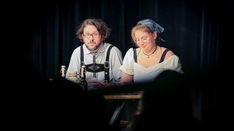 Zwei Personen in traditioneller Kleidung sitzen auf einer Bühne. Sie haben Mikrofone und Getränke vor sich. Der Hintergrund ist ein schwarzer Vorhang., © Theaterhaus Stuttgart e.V. Zwei Personen in traditioneller Kleidung sitzen auf einer Bühne. Sie haben Mikrofone und Getränke vor sich. Der Hintergrund ist ein schwarzer Vorhang., © Theaterhaus Stuttgart e.V.