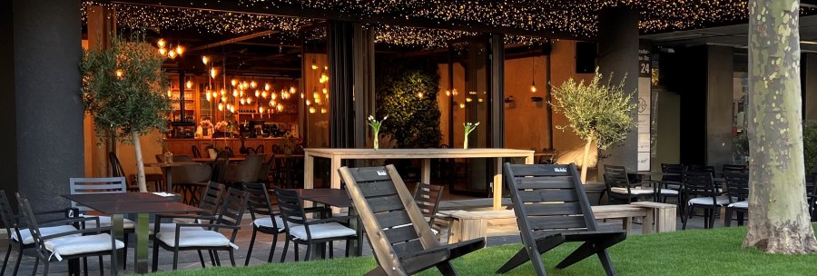 Cozy outdoor area of a café with tables, chairs and fairy lights. The bar can be seen in the background, surrounded by warm light., © heavenskitchen Cozy outdoor area of a café with tables, chairs and fairy lights. The bar can be seen in the background, surrounded by warm light., © heavenskitchen