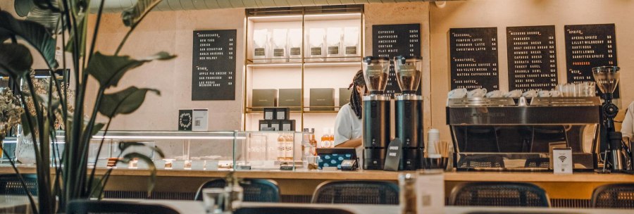Modernes Caf&eacute; mit Theke, Kaffeemaschine und Speisekarten an der Wand. Pflanzen im Vordergrund, gem&uuml;tliche Atmosph&auml;re., &copy; Maison Karl, Stuttgart