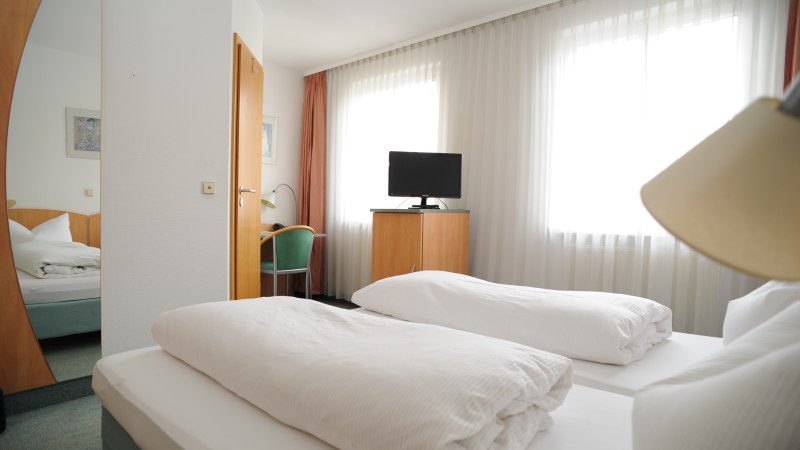 A hotel room with two single beds, a TV on a wardrobe, a desk with chair and large windows with curtains., © TOMAS