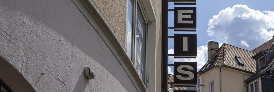 A sign saying 'WHITE' hangs on a building. The sky is blue with a few clouds. Roofs can be seen in the background., © SMG, Sarah Schmid