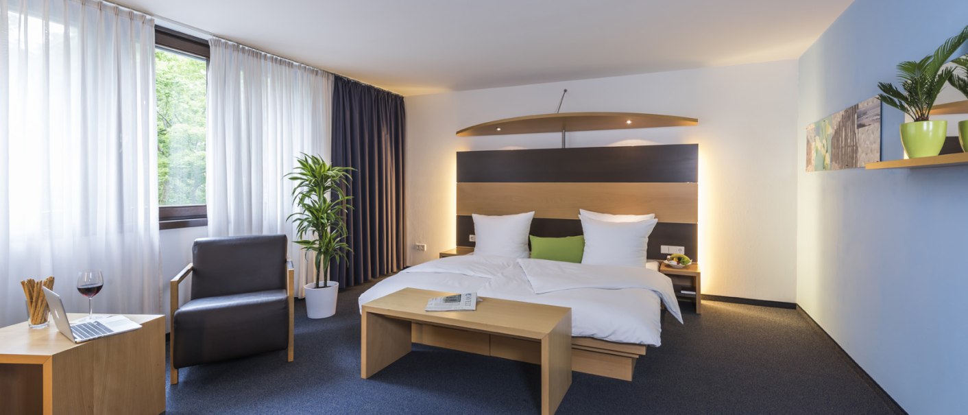 Modernes Doppelzimmer mit großem Bett, Sessel, Tisch mit Laptop und Weinglas, Pflanzen und dekorativen Elementen. Helle und einladende Atmosphäre., © Hotel Berlin aZIS Hotel BetriebsGmbH