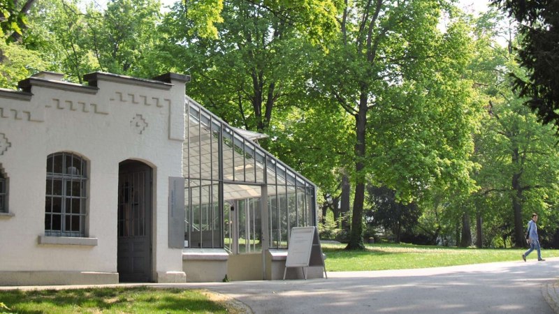 Ein historisches Gebäude mit Glasdach in einem grünen Park. Eine Person geht auf einem Weg entlang., © Stuttgart-Marketing GmbH