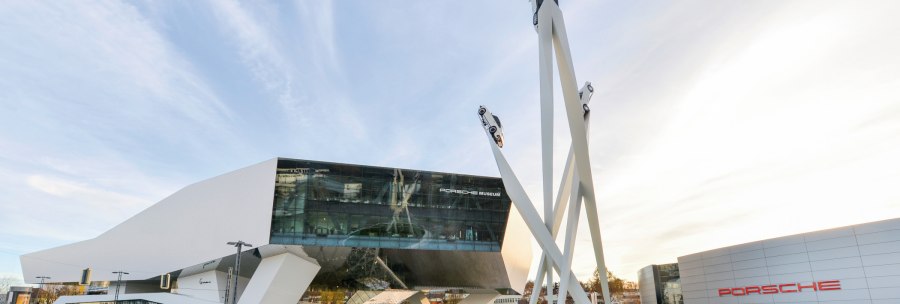 The Porsche Museum with its modern architecture and a sculpture of three cars on high pillars against a blue sky., © Porsche AG, Sabine Braun