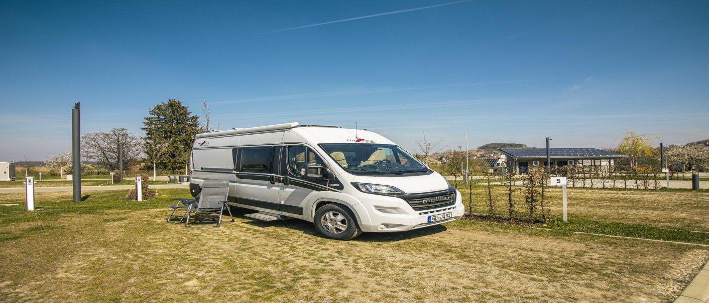 Ein weißes Wohnmobil steht auf einem Stellplatz mit Stühlen davor. Im Hintergrund sind Bäume und ein Gebäude mit Solarpanelen zu sehen., © SMG, Sarah Schmid