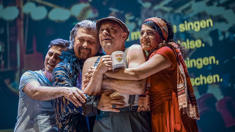 Vier Personen umarmen sich herzlich auf einer Bühne. Eine Person hält einen Becher mit einem Logo. Im Hintergrund sind verschwommene Texte projiziert., © Altes Schauspielhaus, Stuttgart