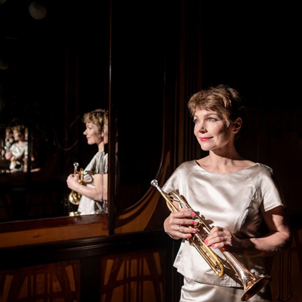 Eine Frau in einem eleganten, hellen Kleid hält eine Trompete und steht vor einem Spiegel, der ihr Bild mehrfach reflektiert., © Altes Schauspielhaus und Komödie im Marquardt e.V.
