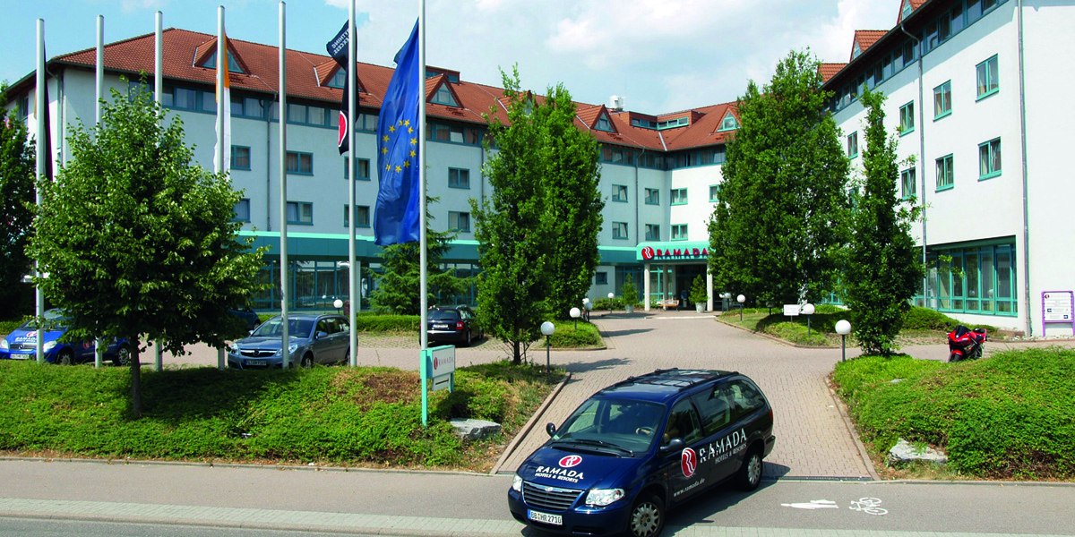 Außenansicht eines Hotels mit Ramada-Logo, umgeben von Bäumen und Flaggen. Ein Ramada-Auto steht im Vordergrund auf der Zufahrt., © © H-Hotels.com Außenansicht eines Hotels mit Ramada-Logo, umgeben von Bäumen und Flaggen. Ein Ramada-Auto steht im Vordergrund auf der Zufahrt., © © H-Hotels.com