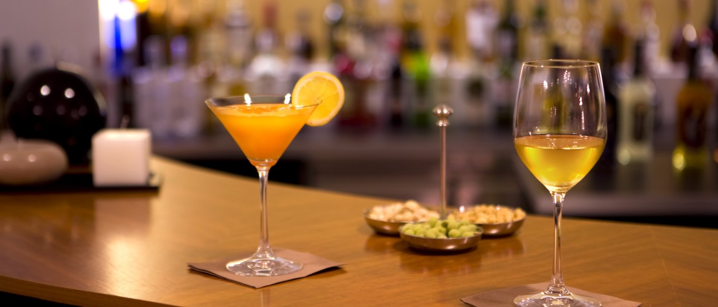 Zwei Cocktails stehen auf einer Hotelbar. Im Hintergrund sind Flaschen und Snacks zu sehen. Die Atmosphäre ist gemütlich und einladend., © Ross Messehotel Zwei Cocktails stehen auf einer Hotelbar. Im Hintergrund sind Flaschen und Snacks zu sehen. Die Atmosphäre ist gemütlich und einladend., © Ross Messehotel