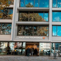 Moderne Glasfassade des Haus des Tourismus mit Spiegelungen von B&auml;umen und blauem Himmel. Menschen betreten die Tourist-Information im Geb&auml;ude, drau&szlig;en stehen Tische und St&uuml;hle., &copy; SMG, Sarah Schmid