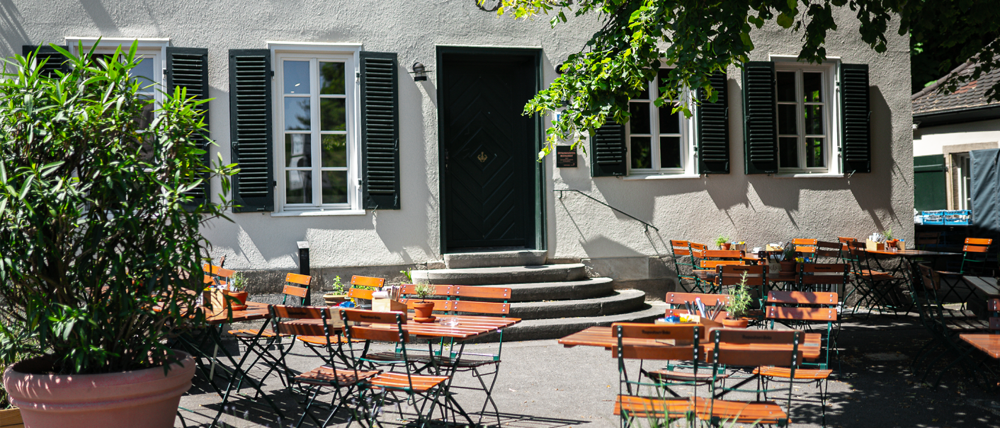 An empty beer garden with wooden tables and chairs in front of a building labeled 'Wirtshaus Hotel'. Shadows from trees fall on the ground., © Cara Schmid An empty beer garden with wooden tables and chairs in front of a building labeled 'Wirtshaus Hotel'. Shadows from trees fall on the ground., © Cara Schmid