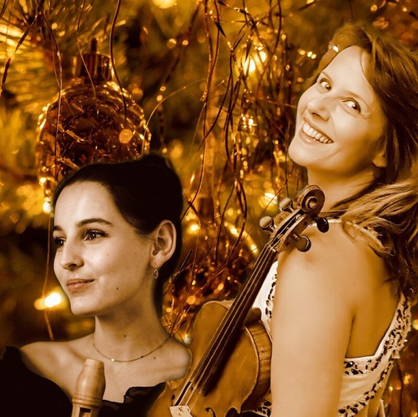 Zwei Frauen mit Geige und Flöte lächeln vor einem festlich geschmückten Weihnachtsbaum. Die Szene ist in warmen, goldenen Tönen gehalten., © Concerti - Costabel