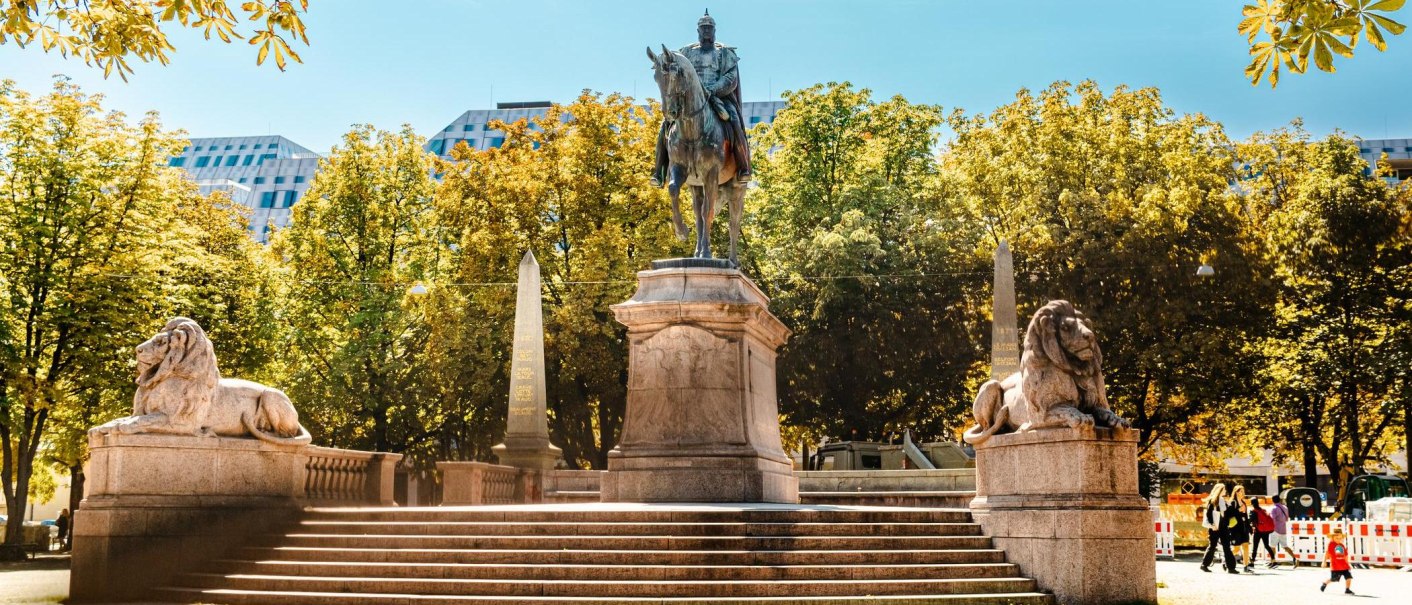 Reiterstatue auf dem Karlsplatz, flankiert von zwei Löwenstatuen. Umgeben von Bäumen und modernen Gebäuden im Hintergrund., © Stuttgart Marketing GmbH, Sarah Schmid