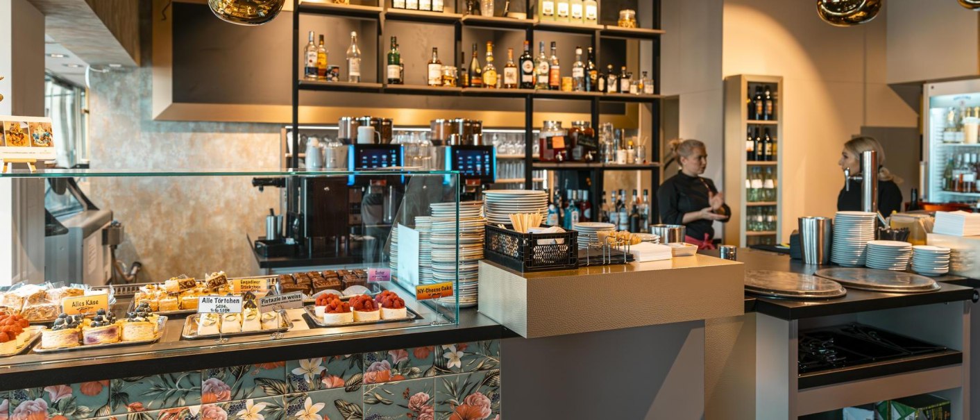 Ein Caf&eacute; mit Theke, Kuchen in einer Vitrine und Regalen mit Flaschen im Hintergrund. Zwei Personen stehen hinter der Theke., &copy; Stuttgart-Marketing GmbH, Sarah Schmid