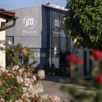 Entrance to the Maier winery, surrounded by flowering plants and trees. Modern architecture with a glass façade and stone walls., © Gottfried Stoppel Entrance to the Maier winery, surrounded by flowering plants and trees. Modern architecture with a glass façade and stone walls., © Gottfried Stoppel