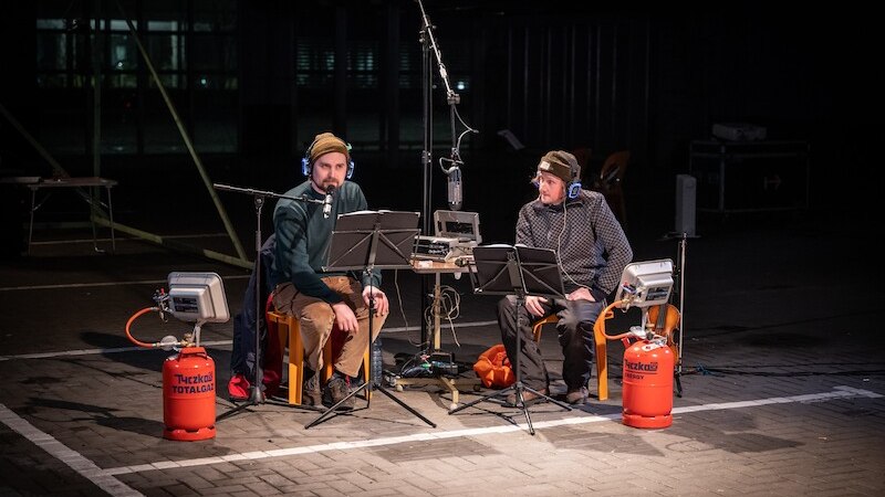 Zwei Männer mit Kopfhörern sitzen an einem Tisch mit Mikrofonen und Notenständern, umgeben von Gasflaschen und Heizgeräten., © Theater Rampe Zwei Männer mit Kopfhörern sitzen an einem Tisch mit Mikrofonen und Notenständern, umgeben von Gasflaschen und Heizgeräten., © Theater Rampe