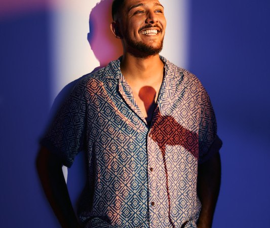 A man in a patterned shirt stands smiling in front of a blue and white illuminated background. The lighting creates interesting shadows., &copy; Rosenau Kultur e.V.