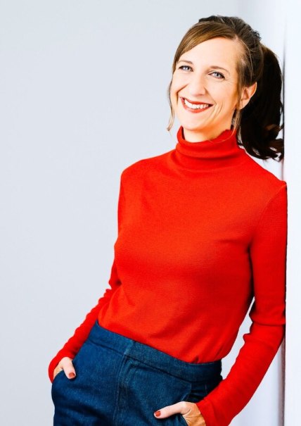 A woman with blonde hair sits smiling at a wooden table in front of a dark background. She is wearing a blue dress., &copy; Rosenau Kultur e.V.