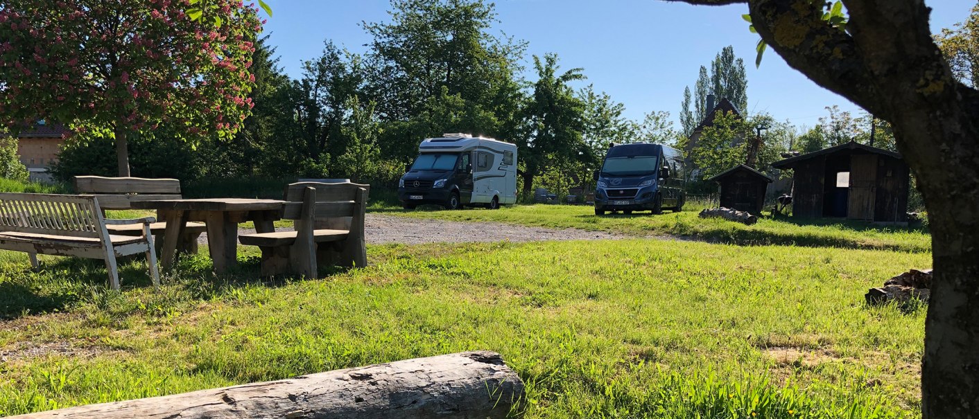 Wohnmobilstellplatz mit zwei Wohnmobilen, Holztisch und B&auml;nken unter einem bl&uuml;henden Baum. Im Hintergrund sind kleine Holzh&uuml;tten zu sehen., &copy; Bio Weingut Zimmer