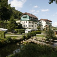 Ein Resort mit Wohnwagen, Gebäuden und einer Holzbrücke in einer grünen, bewaldeten Landschaft unter blauem Himmel., © Family Resort Kleinenzhof Blick in den Nord Schwarzwald
