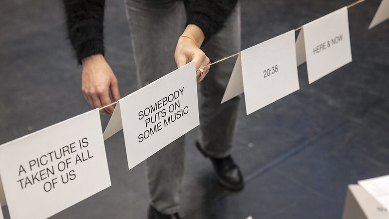 Eine Person hängt Karten mit Texten wie "SOMEBODY PUTS ON SOME MUSIC" an eine Leine. Der Boden ist dunkel, die Person trägt schwarze Schuhe., © Theater Rampe Eine Person hängt Karten mit Texten wie "SOMEBODY PUTS ON SOME MUSIC" an eine Leine. Der Boden ist dunkel, die Person trägt schwarze Schuhe., © Theater Rampe
