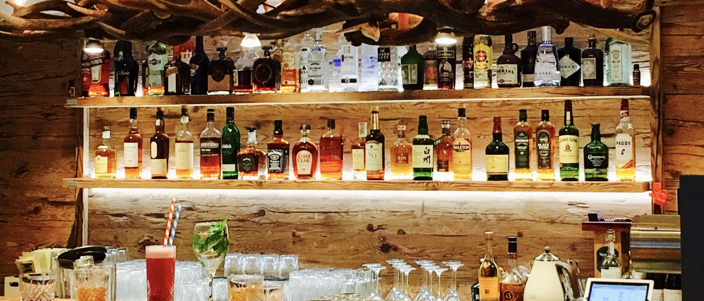 A bar with a wooden counter on which various drinks are placed. Shelves with spirits and an antler lamp can be seen in the background., © TOMAS