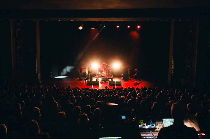 Eine Band spielt auf einer beleuchteten Bühne vor einem großen Publikum in einem dunklen Konzertsaal., © Württembergische Staatstheater Stuttgart