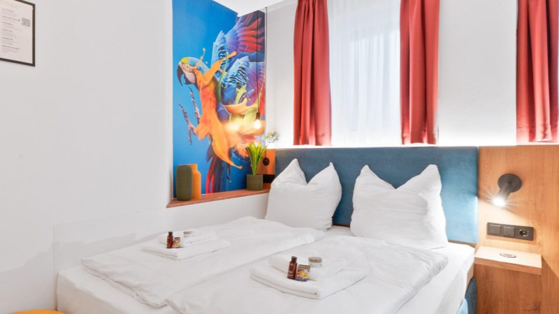 Modernes Doppelzimmer mit weißem Bett, Papageien-Wandbild, roten Vorhängen und gelbem Hocker. Pflegeprodukte auf dem Bett., © JUNGLE N°5 GmbH