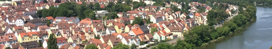 Luftaufnahme einer Stadt mit roten Dächern, einem Fluss und grünen Hügeln im Hintergrund. Der Himmel ist blau mit einigen Wolken., © Aussicht auf den Neckar und die Weinberge Luftaufnahme einer Stadt mit roten Dächern, einem Fluss und grünen Hügeln im Hintergrund. Der Himmel ist blau mit einigen Wolken., © Aussicht auf den Neckar und die Weinberge