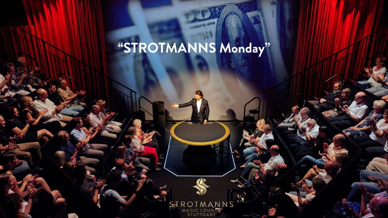 Ein Magier in einem kleinen Theater, umgeben von applaudierendem Publikum. Im Hintergrund sind Geldscheine projiziert. "STROTMANNS Monday" ist zu lesen., &copy; Strotmanns Magic Lounge