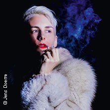 Person mit blondem Haar und Pelzmantel raucht, blauer Rauch im Hintergrund. Dunkler Hintergrund, dramatische Beleuchtung., © links im Bild