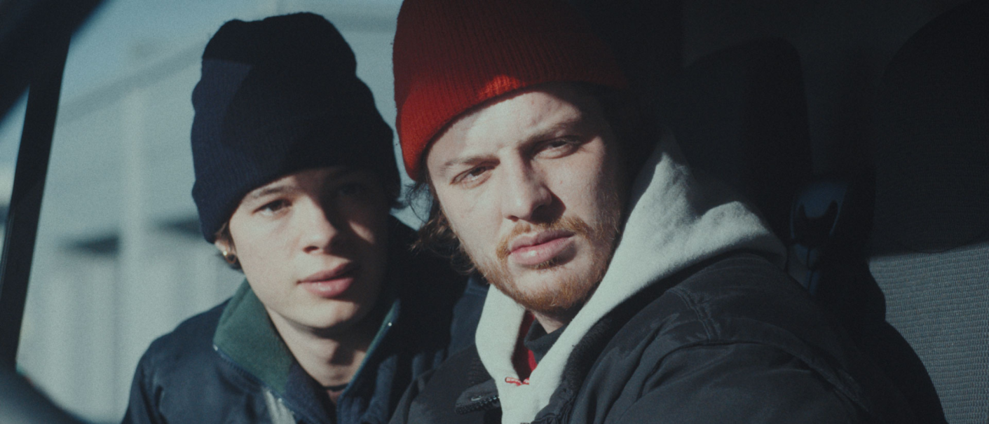 Zwei Männer mit Mützen sitzen in einem Auto. Der vordere trägt eine rote Mütze, der hintere eine blaue. Beide blicken nachdenklich aus dem Fenster., © Pyramide International