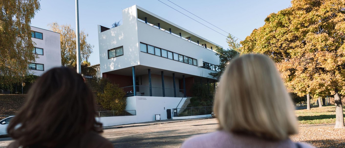 Two people look at the Le Corbusier house in the Weißenhofsiedlung. Modern building with clear lines, surrounded by autumnal trees., © Stuttgart-Marketing GmbH, wpsteinheisser Two people look at the Le Corbusier house in the Weißenhofsiedlung. Modern building with clear lines, surrounded by autumnal trees., © Stuttgart-Marketing GmbH, wpsteinheisser