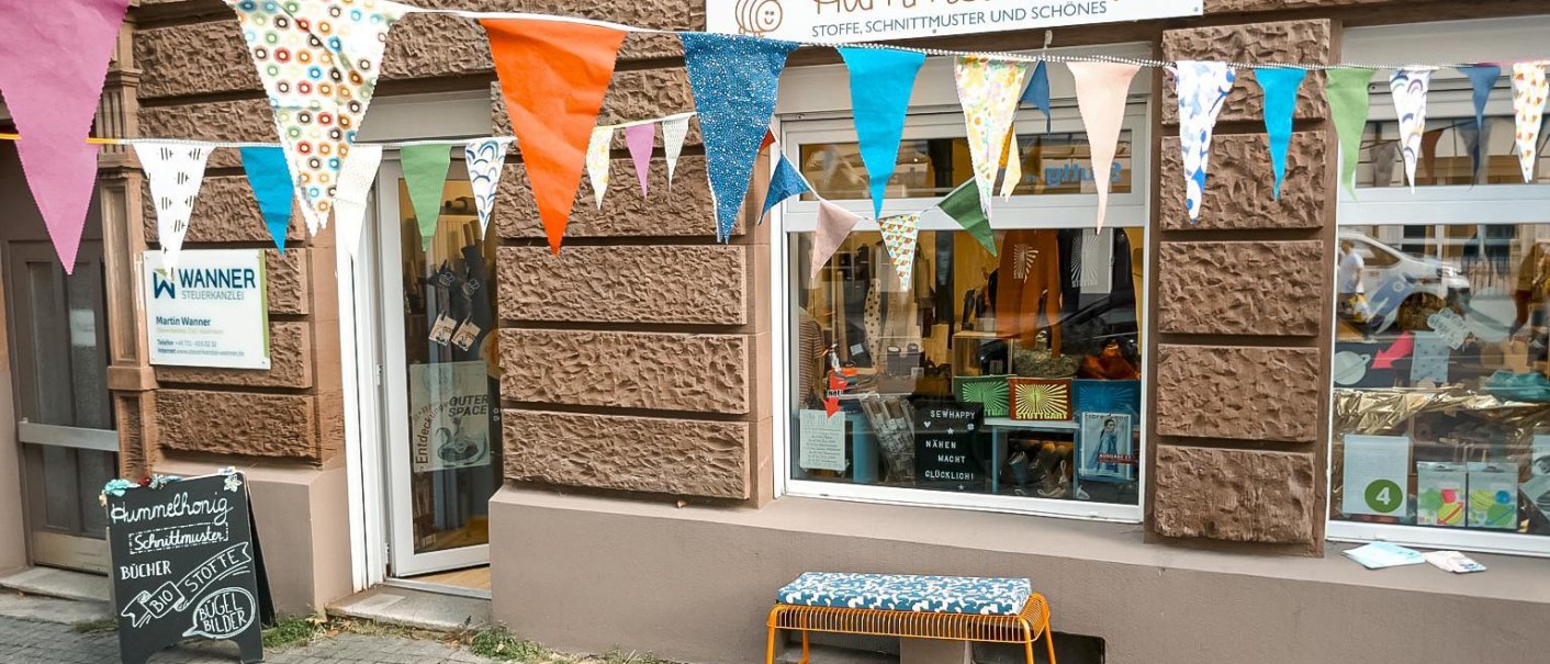 Ladenfront von Hummelhonig mit bunten Wimpeln und einer Bank. Schaufenster zeigt Stoffe und Schnittmuster. Schild der Steuerkanzlei Wanner daneben., © Lena Beck, Hummelkonig GbR, Stuttgart