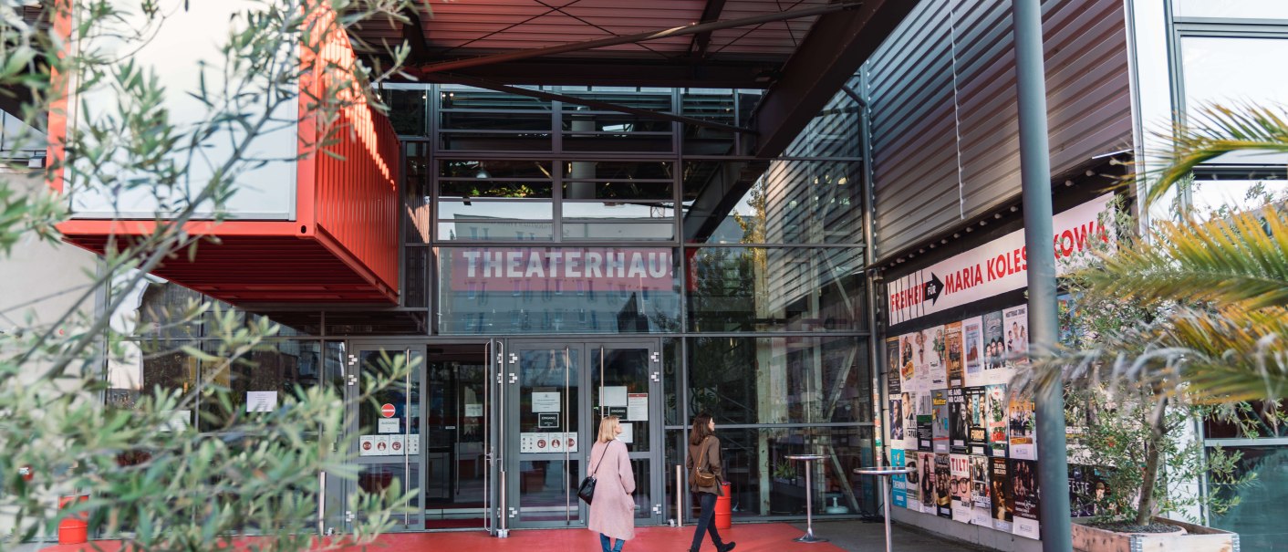 Eingang des Theaterhauses Stuttgart mit rotem Boden, Glasfassade und Plakaten. Zwei Personen betreten das Gebäude. Pflanzen und moderne Architektur sind sichtbar., © Stuttgart-Marketing GmbH, wpsteinheisser