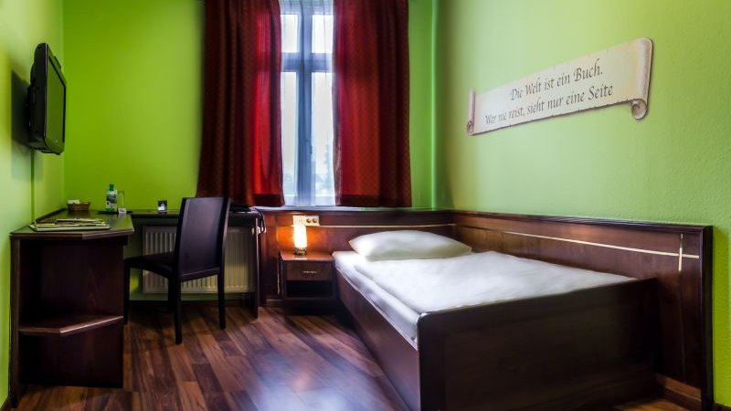 Ein gemütliches Einzelzimmer mit grün gestrichenen Wänden, einem Einzelbett, Schreibtisch und Stuhl. Ein roter Vorhang hängt am Fenster., © TOMAS