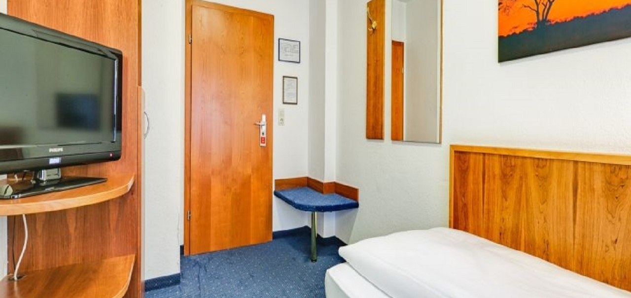Ein gemütliches Einzelzimmer mit einem Bett, einem Fernseher, Holzmöbeln und einem Bild an der Wand. Der Teppich ist blau., © ARNOTEL GmbH