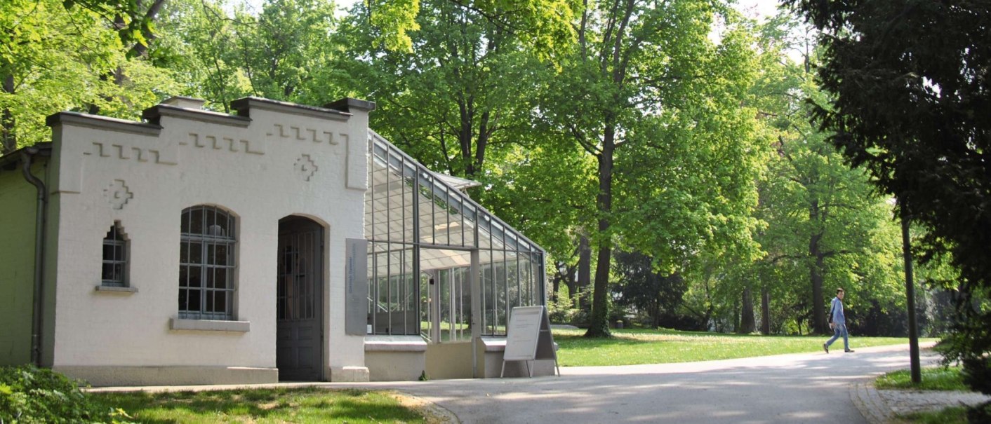Ein historisches Gebäude mit Glasdach in einem grünen Park. Eine Person geht auf einem Weg entlang., © Stuttgart-Marketing GmbH Ein historisches Gebäude mit Glasdach in einem grünen Park. Eine Person geht auf einem Weg entlang., © Stuttgart-Marketing GmbH