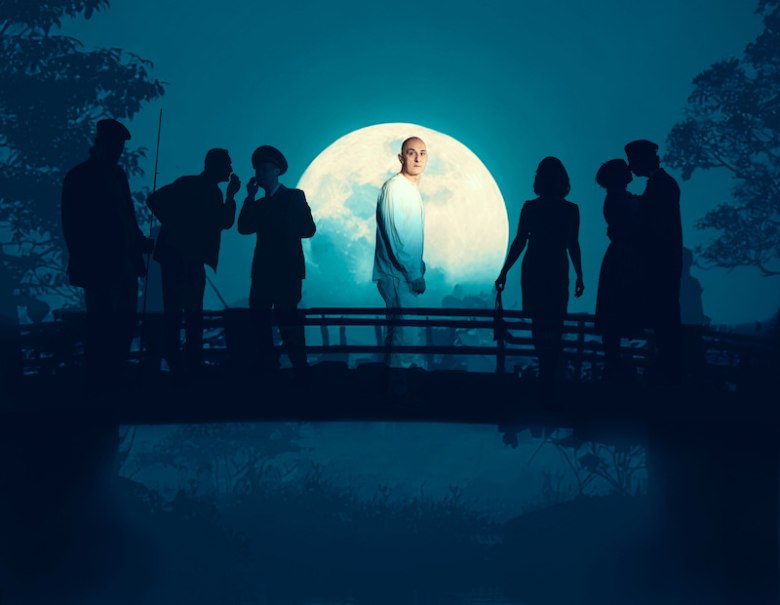 Ein Mann in einem Mantel und Hut sitzt mit ernstem Blick. Das Bild ist in Schwarz-Weiß gehalten., © tri-bühne e.V. Ein Mann in einem Mantel und Hut sitzt mit ernstem Blick. Das Bild ist in Schwarz-Weiß gehalten., © tri-bühne e.V.