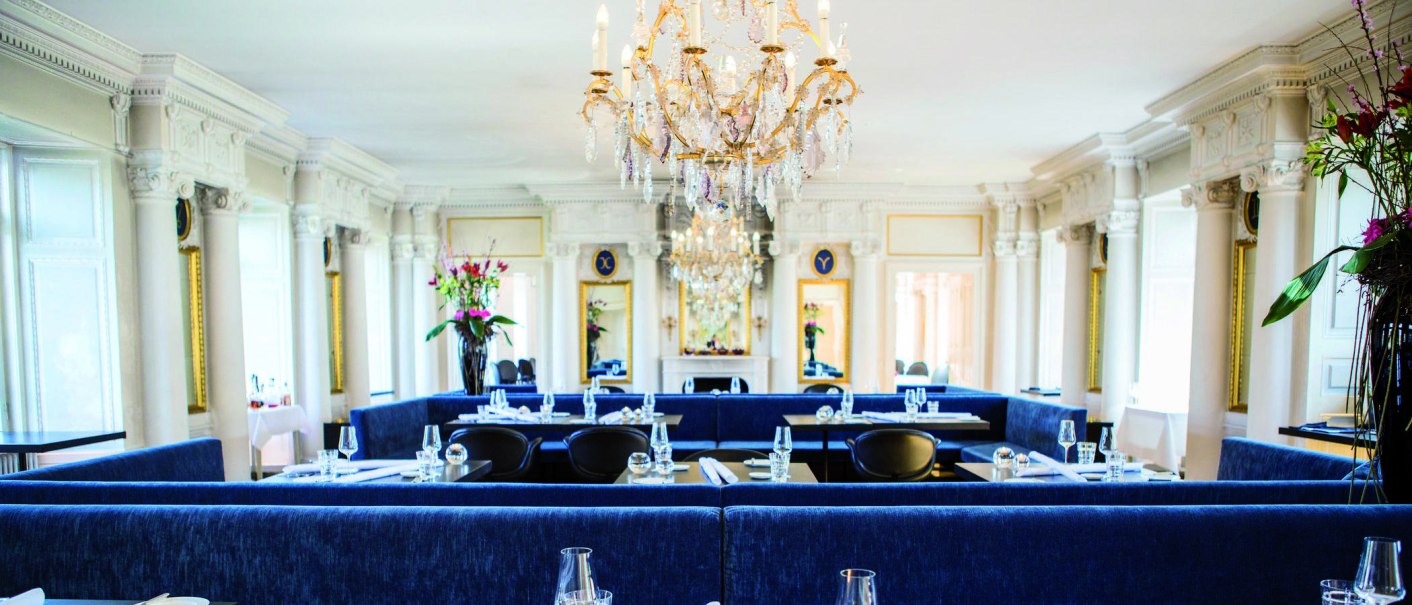 Elegantes Restaurant mit Kronleuchter, blauen Sitzbänken und weißen Säulen. Blumenarrangements und gedeckte Tische schaffen eine luxuriöse Atmosphäre., © Speisemeisterei Elegantes Restaurant mit Kronleuchter, blauen Sitzbänken und weißen Säulen. Blumenarrangements und gedeckte Tische schaffen eine luxuriöse Atmosphäre., © Speisemeisterei