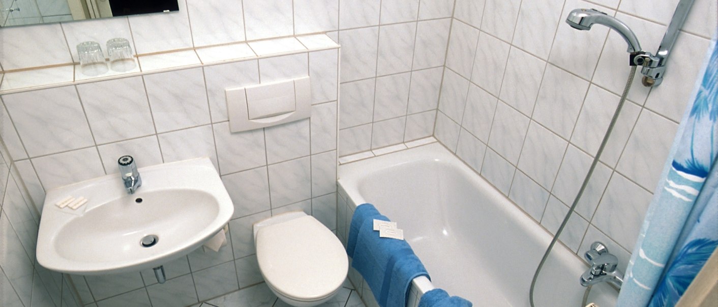 Ein kleines Badezimmer mit weiß gefliesten Wänden, einer Badewanne, einem Waschbecken und einer Toilette. Blaue Handtücher hängen an der Wanne., © Hotel Find