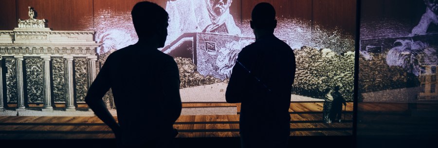 Two people stand in front of a multimedia installation showing Hegel and the Brandenburg Gate. The scene is dark and atmospherically lit., &copy; Julia Ochs